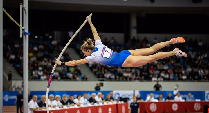 A female pole sport athlete in mid-air, holding a difficult pose during an international competition, with judges visible in the background.